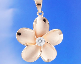 Puede incluir: Un colgante de tono dorado con una flor de plumeria detallada con un centro de cristal transparente.