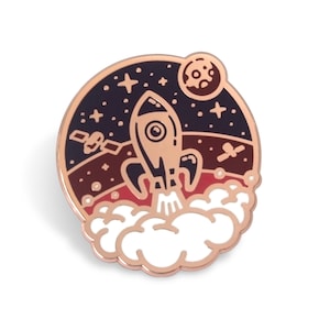 May include: A rose gold enamel pin featuring a rocket ship launching into space. The rocket is white with black details and is surrounded by a dark blue background with white stars and a red planet.