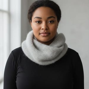 Könnte beinhalten: Ein hellgrauer, gestrickter Loop-Schal. Der weiche, flauschige Schal ist um den Hals gewickelt. Die Person auf dem Bild trägt ein schwarzes Langarmshirt. Der Hintergrund ist ein neutrales Grau.