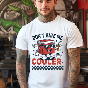 May include: A white short-sleeved t-shirt featuring a graphic of a red cooler wearing sunglasses and holding a drink. The text reads: DON'T HATE ME JUST 'CUZ I'M A LITTLE COOLER, with a black and white checkered pattern printed at the bottom.