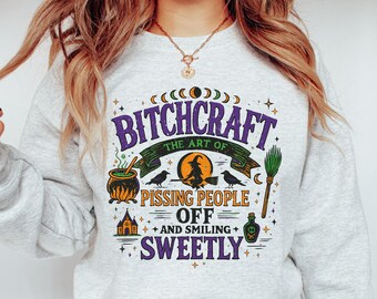 May include: A light grey sweatshirt featuring the phrase "BITCHCRAFT the art of pissing people off and smiling sweetly" in purple, green, and orange. The design includes a witch on a broom, a cauldron, and other witchy symbols.
