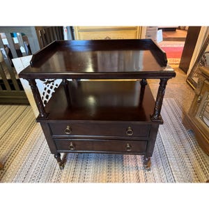 May include: A dark brown wooden side table with two drawers and a top shelf. The table has ornate carved legs and drawer pulls. The top shelf has a raised edge. The table is on a patterned rug.
