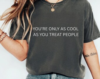 以下が含まれることがあります： 「YOU'RE ONLY AS COOL AS YOU TREAT PEOPLE」の白文字がプリントされた、濃いグレーのTシャツ。クルーネックで、少し色あせたような風合いです。モデルは青いジーンズを着用しています。