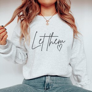 以下が含まれることがあります： 「Let them」という筆記体の文字と小さなハートのシンボルが入った、明るい灰色のスウェットシャツ。柔らかい素材でできており、長袖です。モデルは青いジーンズを着用しています。