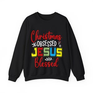 Puede incluir: Sudadera negra con las palabras "Christmas OBSESSED JESUS Blessed" en rojo, amarillo, verde y azul. La sudadera tiene cuello redondo y mangas largas. El texto está en una fuente festiva y colorida.
