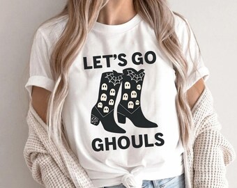 Puede incluir: Camiseta blanca con el texto "LET'S GO GHOULS" y un gráfico de botas de vaquero negras decoradas con telarañas y diseños de calaveras. Una prenda casual para el día a día.
