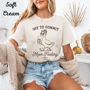 May include: A cream-colored short-sleeve t-shirt featuring a brown graphic of a duck wearing a headband and heart-shaped sunglasses while holding a coffee cup. The text reads OFF TO COMMIT Mom Foolery. The shirt is paired with blue denim shorts.