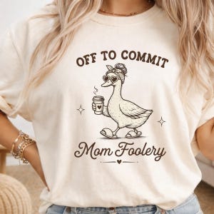 May include: A cream-colored t-shirt featuring a brown graphic of a duck wearing a bandana, heart-shaped sunglasses, and slippers while holding a coffee cup. The text reads OFF TO COMMIT Mom Foolery with small star and heart accents around the duck.