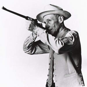 May include: A man wearing a white hat and waistcoat holds a rifle and aims it at the camera.
