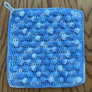 Pode incluir: Um pegador de panela quadrado em crochê com um padrão texturizado de pontos pipoca em vários tons de azul e branco. O pegador possui uma pequena alça no canto superior esquerdo para pendurar e um design de fio denso e repetitivo.