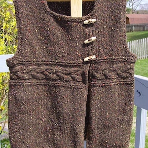 Op de afbeelding: Een bruin gebreid vest met een kabelbreipatroon en een knoopsluiting. Het vest heeft een gespikkeld patroon en is gemaakt van een zacht, warm garen.