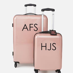 May include: Two pink suitcases with black handles and wheels. The larger suitcase has the initials "AFS" printed on the front. The smaller suitcase has the initials "HJS" printed on the front and a luggage tag that says "Take me with you!"