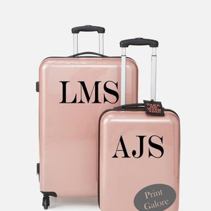 May include: Two pink suitcases with black handles and wheels. The larger suitcase has the initials "LMS" printed on it. The smaller suitcase has the initials "AJS" printed on it and a sticker that says "Take me with you!" and "Print Galore".