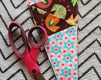 Könnte beinhalten: Eine rosa Schere liegt auf einem gemusterten Stoff mit einem passenden Scherenhalter. Der Halter hat ein Vogel- und Blumendesign in Braun-, Rosa- und Blautönen. Die Schere ist etwa 20 cm lang.