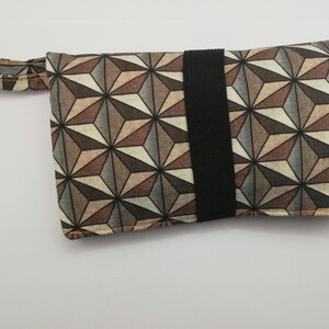 Puede incluir: Una funda rectangular con un patrón geométrico en tonos marrón, gris y beige. Cuenta con una banda elástica negra y un mosquetón dorado sujeto a un lazo de tela. La funda está diseñada para objetos pequeños.