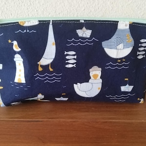 Può includere: Pochette con cerniera blu navy con un motivo nautico con gabbiani, barche a vela, fari e pesci. La pochette ha una cerniera verde menta e un piccolo anello di tessuto. Una conchiglia poggia sulla superficie di legno.