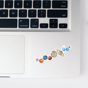 K&ouml;nnte beinhalten: Ein Aufkleber des Sonnensystems mit den Planeten in der Reihenfolge von der Sonne aus. Die Planeten sind in Aquarell gemalt und befinden sich in einer Linie auf einem silbernen Laptop.