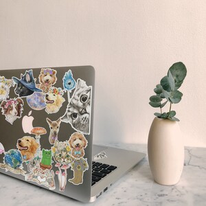 May include: A laptop computer with a white background and a variety of colorful animal stickers on the lid. The stickers feature cats, dogs, and other animals. A vase with a green plant is on the right side of the image.