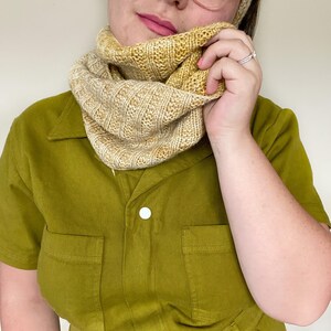 May include: A person wearing a yellow and white knit cowl neck scarf. The scarf is folded over and worn around the neck. The person is wearing a green collared shirt.