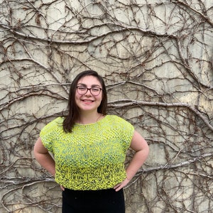 Puede incluir: Una persona lleva un top corto de punto amarillo y verde con una falda negra. La persona est&aacute; de pie frente a una pared cubierta de enredaderas.
