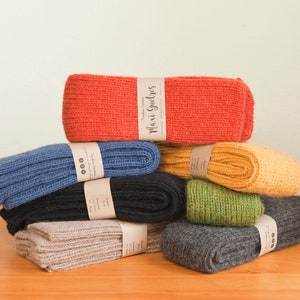 May include: A stack of eight pairs of knitted leg warmers in various colors, including red, blue, black, beige, green, and yellow. The leg warmers are folded and stacked on a wooden surface. Each pair has a small paper tag with the text "Maxi Gürtels" on it.