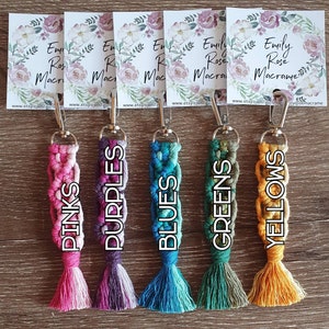 May include: Five colorful macrame keychains with tassels. The keychains are pink, purple, blue, green, and yellow. Each keychain has a silver clasp and is labeled with the color name.