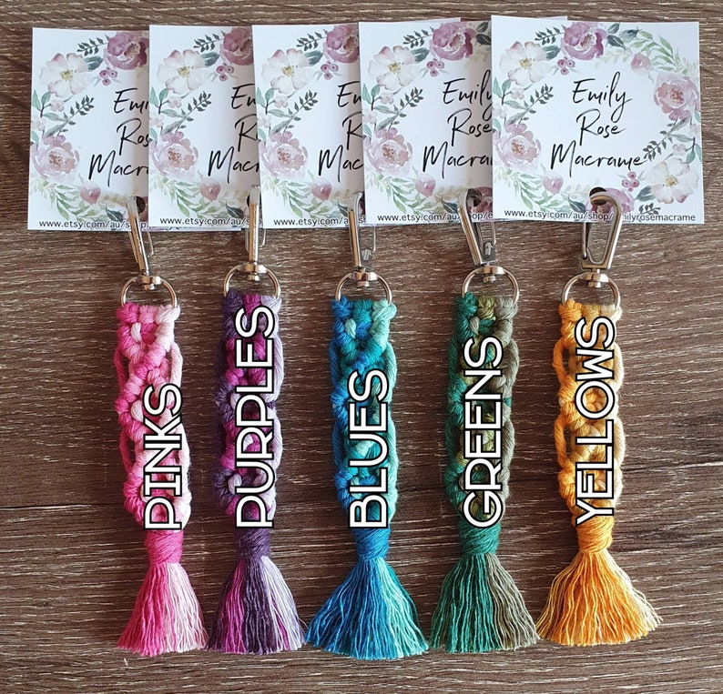 May include: Five colorful macrame keychains with tassels. The keychains are pink, purple, blue, green, and yellow. Each keychain has a silver clasp and is labeled with the color name.