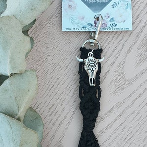 May include: Black macrame keychain with a silver bull skull charm. The keychain has a black tassel and a silver key ring. The keychain is on a wooden surface with a floral card behind it. The card says "Emily Rose Macrame" and "www.etsy.com/au/shop/emilyrosemacrame".