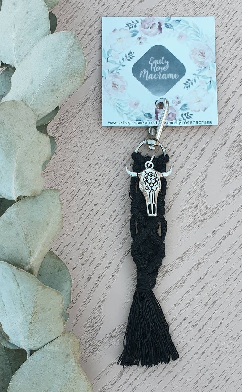 May include: Black macrame keychain with a silver bull skull charm. The keychain has a black tassel and a silver key ring. The keychain is on a wooden surface with a floral card behind it. The card says "Emily Rose Macrame" and "www.etsy.com/au/shop/emilyrosemacrame".