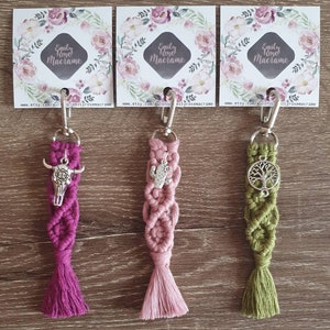 May include: Three macrame keychains with different charms. The keychain on the left has a pink tassel and a silver skull charm. The middle keychain has a pink tassel and a silver cactus charm. The keychain on the right has a green tassel and a silver tree charm. Each keychain has a silver metal ring and a silver clasp.