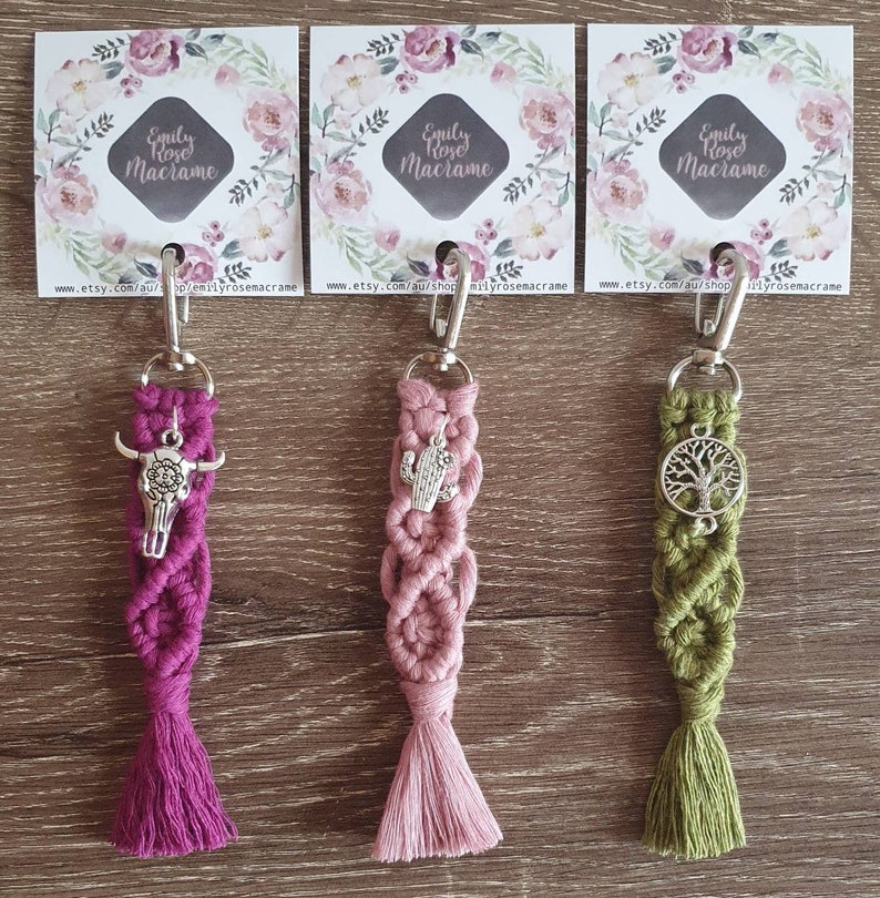 May include: Three macrame keychains with different charms. The keychain on the left has a pink tassel and a silver skull charm. The middle keychain has a pink tassel and a silver cactus charm. The keychain on the right has a green tassel and a silver tree charm. Each keychain has a silver metal ring and a silver clasp.