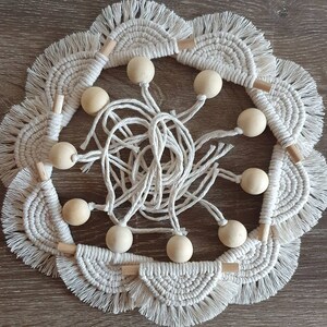 May include: A white macrame wall hanging with wooden beads. The macrame is made of cotton cord and has a fringe border. The beads are natural wood and are strung on the macrame.
