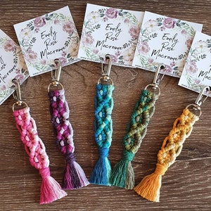 May include: Five colorful macrame keychains with silver key rings and tassels. The keychains are made with different colored cords and have a braided design. Each keychain has a small card attached with the text "Emily Rose Macrame" and a floral design.