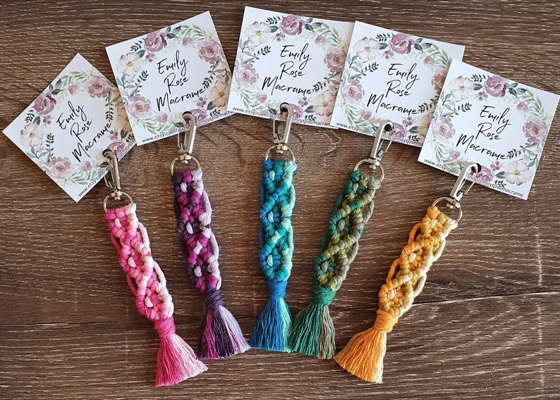 May include: Five colorful macrame keychains with silver key rings and tassels. The keychains are made with different colored cords and have a braided design. Each keychain has a small card attached with the text "Emily Rose Macrame" and a floral design.