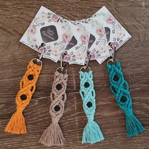 May include: Four colorful macrame keychains with black lava rock beads and tassels. The keychains are displayed on a wooden surface with business cards featuring the text "Emily Rose Macrame" and a website address.