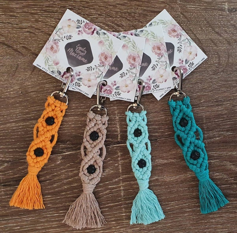 May include: Four colorful macrame keychains with black lava rock beads and tassels. The keychains are displayed on a wooden surface with business cards featuring the text "Emily Rose Macrame" and a website address.