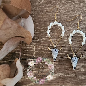 May include: A pair of white macrame earrings with a gold-tone bull skull charm. The earrings are hanging from a wooden surface.