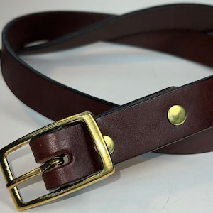 May include: A dark brown leather belt with a gold-colored rectangular buckle and two gold-colored rivets. The belt is curved, showing its flexibility. The buckle is a classic design.