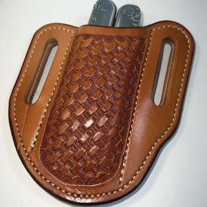 May include: A brown leather multi-tool sheath with a woven pattern on the front. The sheath has two slots for a belt and holds a silver multi-tool. The edges are stitched with white thread.