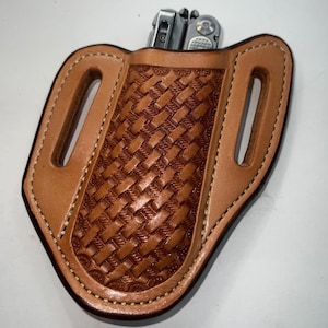 May include: A light brown leather multi-tool sheath with a woven basket pattern. The sheath has a stitched border and belt loops. A silver multi-tool is partially visible in the sheath.