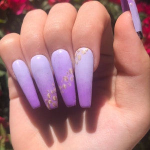 May include: Long, light purple ombre press-on nails with gold glitter accents.