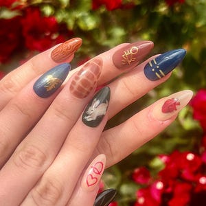 May include: Close-up of a hand with long, almond-shaped nails painted with various designs. The nails feature a brown and white animal print, a black nail with a white rabbit, a brown nail with a gold key, a blue nail with gold stripes, a nude nail with a red heart, and a black nail with a gold accent.