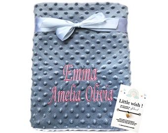 May include: A gray, soft, plush baby blanket with a white satin ribbon tied in a bow. The blanket is personalized with the names "Emma, Amelia, Olivia" embroidered in pink. A small card with a rainbow and the text "Little wish! Jewel" is included.
