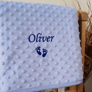 May include: A light blue baby blanket with a white border. The blanket is embroidered with the name "Oliver" and a pair of footprints with hearts.