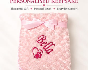 May include: A soft, light pink textured blanket featuring a satin ribbon tied in a bow at the top. The blanket is embroidered with the name Bella in dark pink script, accompanied by a dark pink heart icon containing a small paw print design.