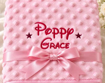 May include: A soft pink dimpled minky blanket featuring the embroidered name Poppy Grace in dark pink thread, accented by two small dark pink stars on either side of the name, and finished with a matching pink satin ribbon tied in a bow.