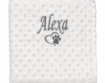 May include: A white square blanket with a raised dimpled texture. The center features grey embroidery of the name Alexa in a cursive script font, positioned above a grey heart outline that incorporates a paw print design at the bottom.