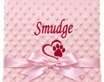 May include: A light pink dimpled minky blanket featuring the embroidered name Smudge in dark pink script above a dark pink heart and paw print design. A matching light pink satin ribbon is tied in a bow across the lower section of the blanket.