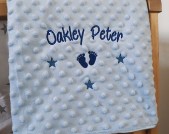 May include: A light blue baby blanket with a textured, dotted surface. The blanket features the embroidered name "Oakley Peter" in navy blue, along with navy blue footprints and star designs. The blanket is draped over a wooden crib.