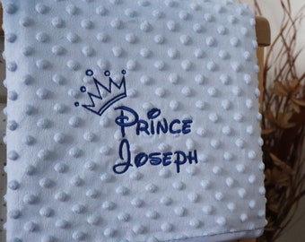 May include: A light blue baby blanket with a textured, dotted surface. The blanket features an embroidered crown and the name "Prince Joseph" in dark blue thread. The blanket is folded over a wooden crib.
